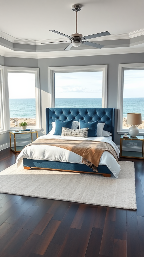 Modern coastal bedroom with a blue headboard, large windows overlooking the ocean, and warm wood flooring.